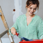 Young woman wearing green striped shirt paints a room during a home renovation that would increase her home insurance coverage needs.