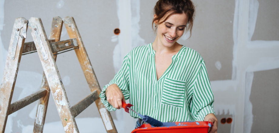 Young woman wearing green striped shirt paints a room during a home renovation that would increase her home insurance coverage needs.