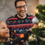 Father and daughter wearing holiday sweaters decorate a pine Christmas tree with silver and gold ornaments.