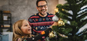 Father and daughter wearing holiday sweaters decorate a pine Christmas tree with silver and gold ornaments.