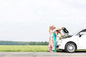 Two female friends examine a broken-down white car stalled from heat-related issues in summer.