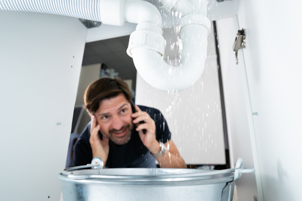 Worried middle-aged man uses his cell phone to call a plumber about a leaking pipe that may be covered by his homeowners insurance.
